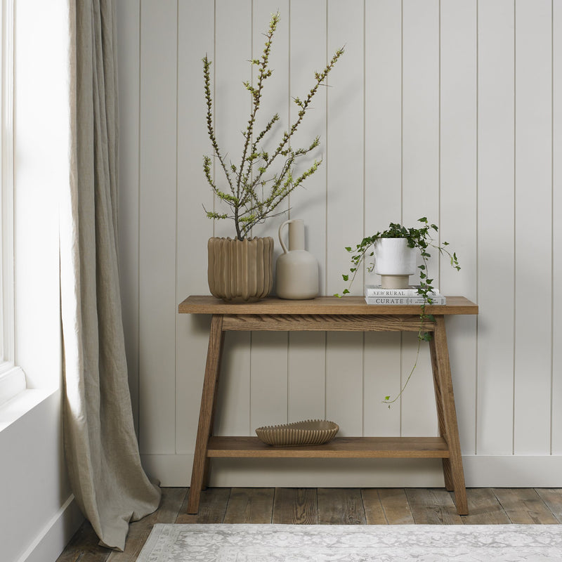 The Camden Console Table with oak tops stands against a light paneled wall, holding three vases with branches and greenery. A ribbed bowl is on the lower shelf, with beige curtains and a patterned rug partially visible.
