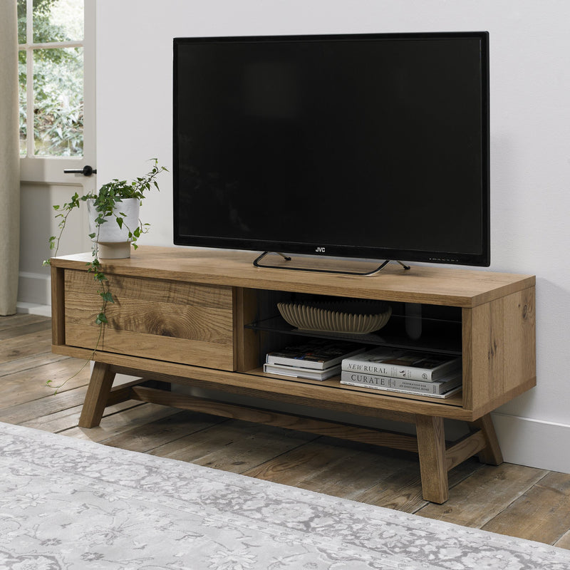 A flat-screen TV is placed on the Camden TV Unit, which features angled legs and mid-century style. The unit has an open shelf with books and a bowl, a closed cabinet, and a potted plant on top. Nearby are a window and a light rug.