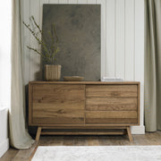 The Camden Large Sideboard features two doors, two drawers, and angled legs in a mid-century modern style. On top, there’s a vase with green branches, books, and a large gray artwork set against a white paneled wall.