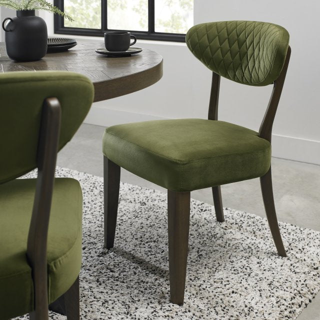 The Ellipse Fumed Oak Upholstered Chair with green velvet fabric and dark wooden legs is placed next to a rustic oak dining table on a speckled rug in a bright, minimalistic room with large windows.