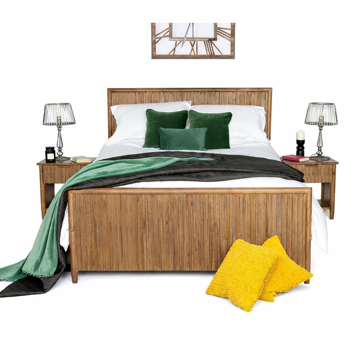 The Cairo Bedframe from the rustic bedroom collection features white bedding, green pillows, and a black and green throw. Bedside tables with lamps, two yellow cushions, and a wall clock complete this sustainable furniture setup.