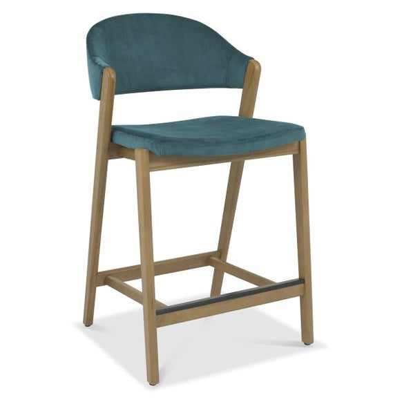 The Camden Counter Stool features light wooden legs, a teal upholstered seat and backrest, a low curved retro-inspired back, and a footrest across the front two legs.