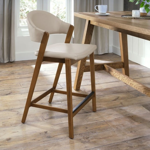 The Camden Counter Stool, featuring a modern wooden frame, light beige cushioned seat, and curved back, stands by a wooden dining table in a bright room with wood floors and large windows. A cup and saucer rest on the table. Ideal for fans of curved back stools.