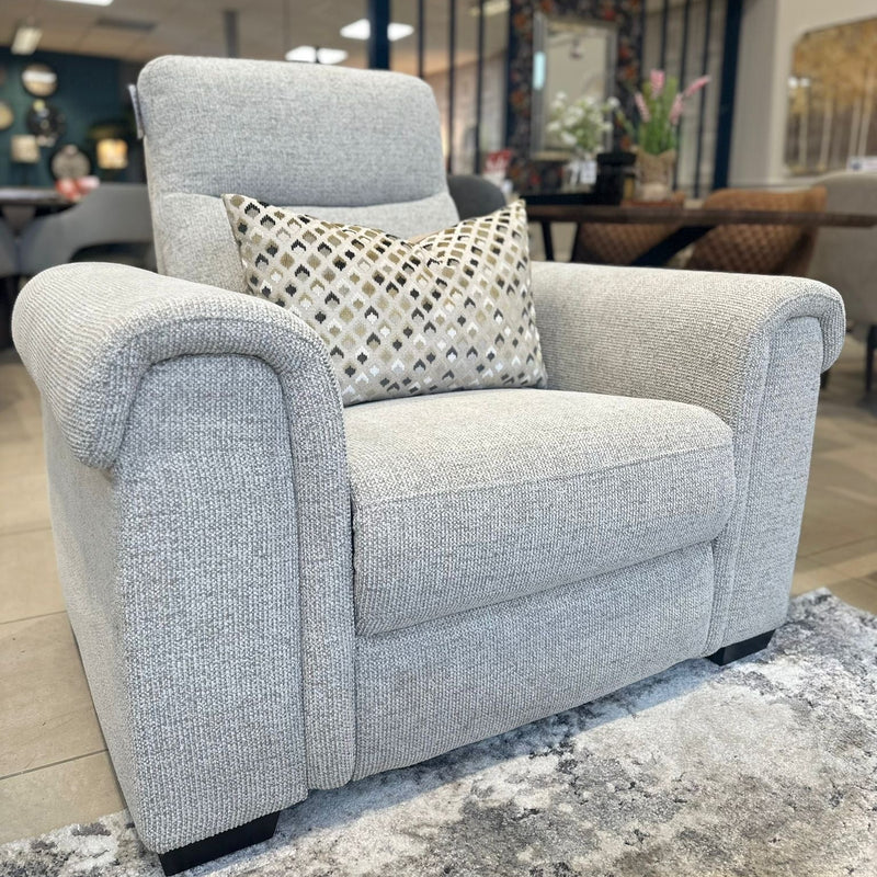 A light gray upholstered armchair with a geometric cushion sits on a gray rug in a showroom, next to a modern sofa and the Luna Electric Recliner, with other furniture and decor visible in the blurred background.