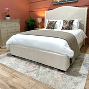 A modern bedroom featuring the Venice Bedframe 5ft—a cream upholstered bedframe with a curved headboard—paired with white and taupe bedding, two patterned pillows, a gray dresser, potted plants, a lamp, and a light patterned rug atop wooden floors.