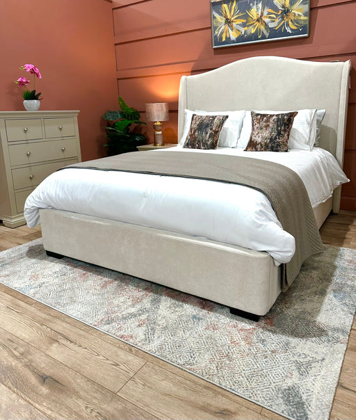 A cozy bedroom featuring the Venice Bedframe upholstered in cream, white bedding, two decorative pillows, a taupe throw, a dresser, potted plants, warm lighting, and floral artwork on a terracotta accent wall.