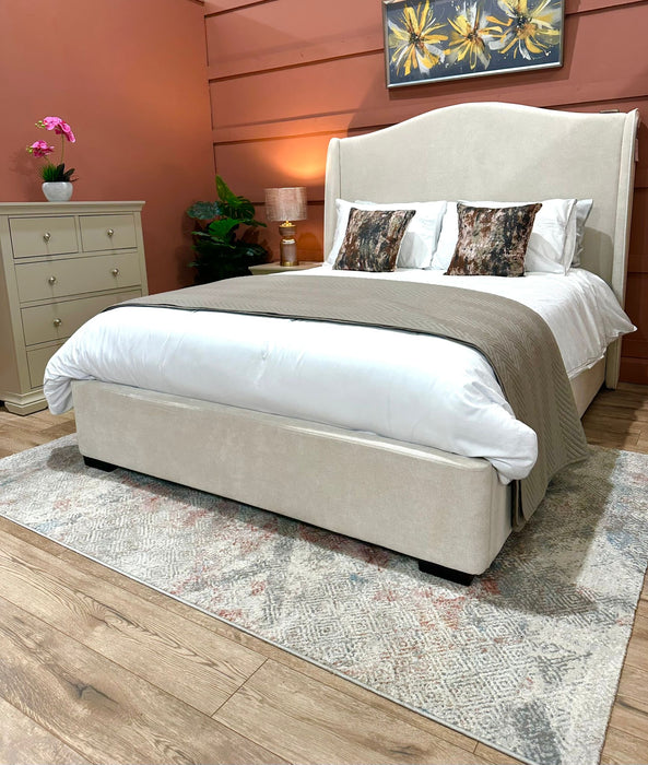 A cozy bedroom featuring the Venice Bedframe upholstered in cream, white bedding, two decorative pillows, a taupe throw, a dresser, potted plants, warm lighting, and floral artwork on a terracotta accent wall.