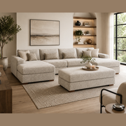 A modern living room featuring the Henley Sofa in beige chenille fabric, a matching ottoman, textured rug, large window, potted tree, wall shelves with decor, and a neutral abstract painting.
