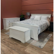 A white wooden bedframe with a high headboard and polished chrome cup handles, displayed in a bedroom setting with bedding and a nightstand.