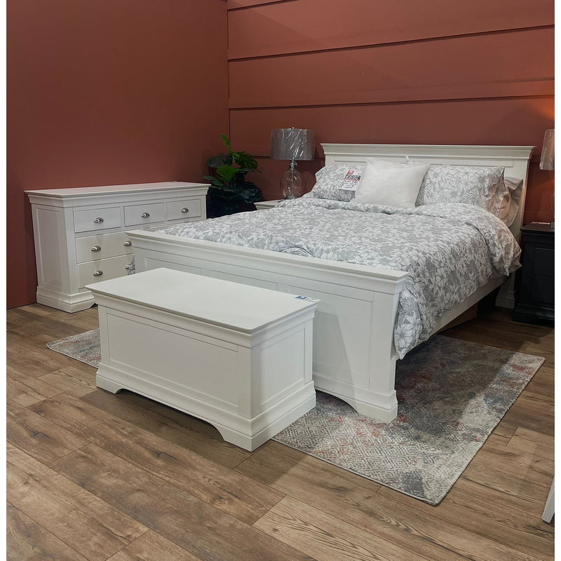 A white wooden bedframe with a high headboard and polished chrome cup handles, displayed in a bedroom setting with bedding and a nightstand.