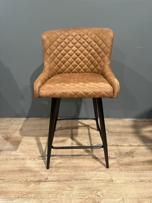 The Diamond Barstool features brown PU leather with quilted diamond stitching, a curved backrest, and black metal legs. Shown on a wooden floor by a gray wall, it’s ideal for your kitchen island.