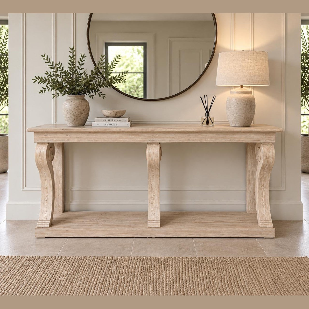 A Montclair Console Table is elegantly set against a paneled wall above a woven rug, styled with a vase of greenery, books, a round mirror, a lamp, and reed diffusers.