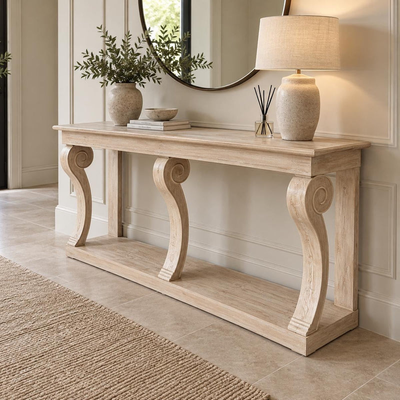 A light Montclair Console Table with ornate legs is styled with a lamp, book, bowl, and reed diffuser. A round mirror and potted plants accent the neutral, elegant hallway featuring tile flooring.