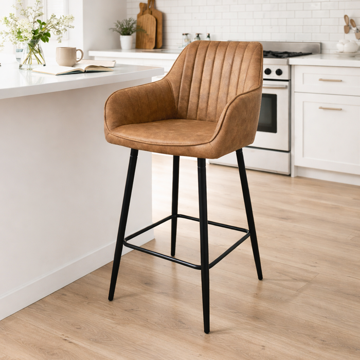 A modern kitchen with light wood floors, white cabinets, and a white countertop features the Enzo Counter Stool—tan leather with black metal legs—at the island. Plants and kitchenware decorate the background.