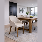 A modern dining room featuring a wooden table, Como Dining Chair in light fabric, floral vase, apple bowl, neutral rug, and large window with city view. Artwork and black cabinets appear in the background.