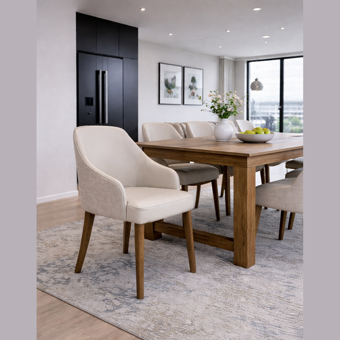 A modern dining room featuring a wooden table, Como Dining Chair in light fabric, floral vase, apple bowl, neutral rug, and large window with city view. Artwork and black cabinets appear in the background.