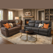 A cozy living room with a comfortable Logan Recliner in brown and gray, colorful cushions, a round coffee table with a Hermes book and chocolate, a stone fireplace, shelves, and large windows with beige curtains.
