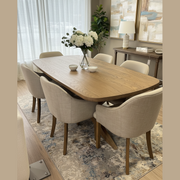 A modern dining room features the Ashton Dining Table in solid ash wood, surrounded by six upholstered chairs. A vase with white flowers sits atop the table, complemented by abstract art, a lamp, and a potted plant in a bright space.