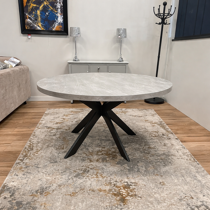 The Rimini Round Extending Table with a gray stone top and black metal legs stands on a beige patterned rug in a living room featuring wood floors, neutral walls, and contemporary decor.