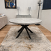 The Rimini Round Extending Table with a gray stone top and black metal legs stands on a beige patterned rug in a living room featuring wood floors, neutral walls, and contemporary decor.