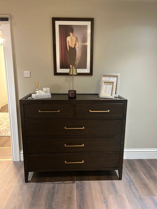 The Hampshire Tall Chest with a dark wood finish and gold handles stands against a beige wall, topped with framed photos, a small lamp, and a diffuser. Above it, artwork depicts a woman in a black dress with her back turned.