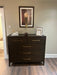 The Hampshire Tall Chest with a dark wood finish and gold handles stands against a beige wall, topped with framed photos, a small lamp, and a diffuser. Above it, artwork depicts a woman in a black dress with her back turned.