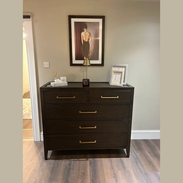The Hampshire Tall Chest with a dark wood finish and gold handles stands against a beige wall, topped with framed photos, a small lamp, and a diffuser. Above it, artwork depicts a woman in a black dress with her back turned.