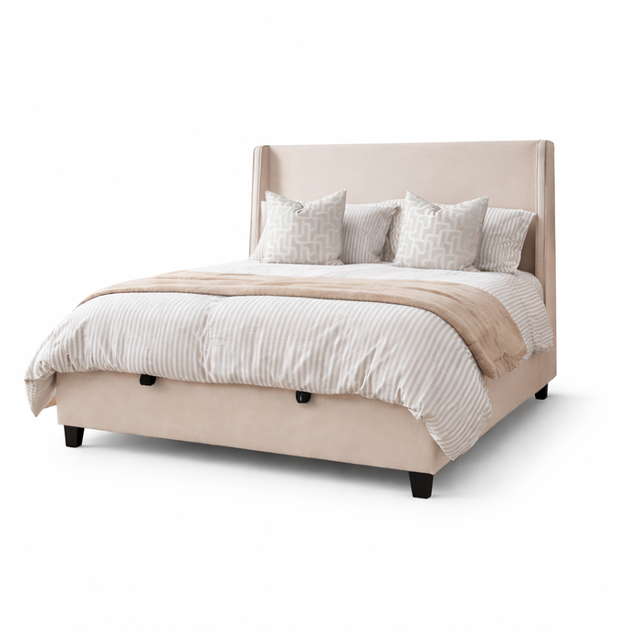 The Rivera Storage Bedframe, upholstered in modern beige linen with a tall headboard, is styled with white and beige striped bedding, four patterned pillows, and a beige throw blanket set against a white background.