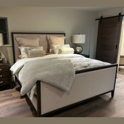 A modern bedroom featuring the Hampshire Bedframe in dark wood, beige and silver bedding, two globe lamps on matching nightstands, a gray throw blanket, a sliding wooden door, wood flooring, and neutral-colored walls.