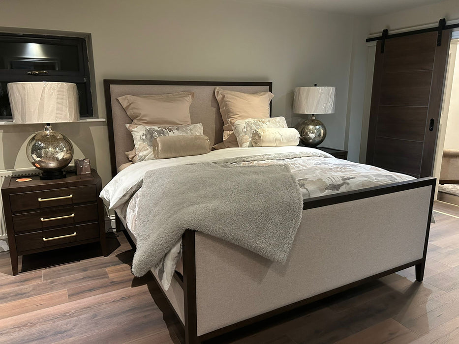 A modern bedroom featuring the Hampshire Bedframe in dark wood, beige and silver bedding, two globe lamps on matching nightstands, a gray throw blanket, a sliding wooden door, wood flooring, and neutral-colored walls.