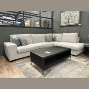 The Sorrento Chaise Corner - Special Offer, upholstered in light gray neutral-textured fabric with spacious seating, is adorned with patterned accent pillows. Positioned next to a dark wood coffee table atop a light rug in a modern showroom, it’s set against gray walls featuring curated artwork and décor.