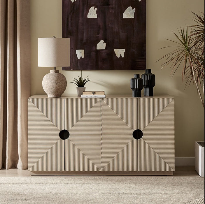 The Arbor 4 Door Sideboard, featuring a bold geometric design and spacious storage, is accented with a textured lamp, two dark vases, and a small plant. Above it hangs abstract art beside beige curtains and a tall potted plant.
