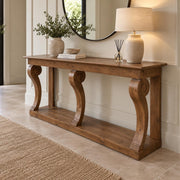 The Montclair Console Table is elegantly styled with ornate curved legs and adorned with a beige lamp, a vase filled with greenery, stacked books, and a reed diffuser. A mirror and potted plant in the background enhance the sense of depth, while a tan rug lies atop the tiled floor.