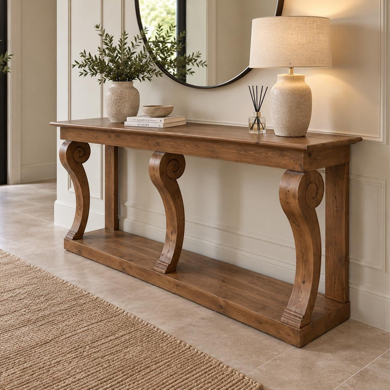 The Montclair Console Table is elegantly styled with ornate curved legs and adorned with a beige lamp, a vase filled with greenery, stacked books, and a reed diffuser. A mirror and potted plant in the background enhance the sense of depth, while a tan rug lies atop the tiled floor.
