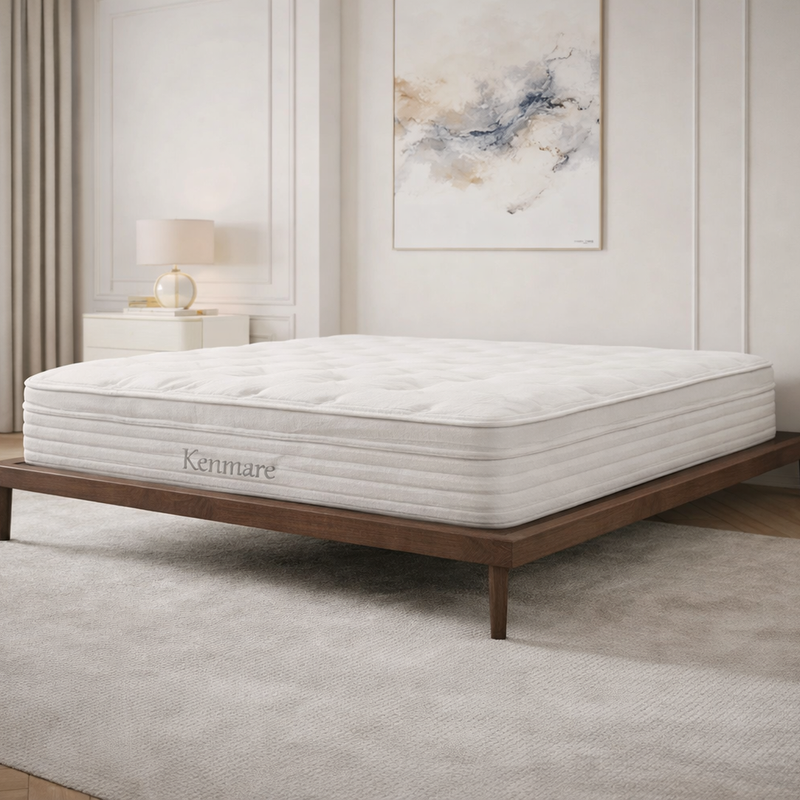 A modern bedroom features a spacious Kenmare Mattress with edge-to-edge support on a low wooden bed frame, light curtains, a rug, a nightstand with lamp, dresser, and abstract neutral wall art.
