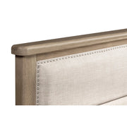 A close-up of the Aylesbury bedframe, showcasing its oak frame and light beige upholstered headboard, accented with a row of decorative silver nailhead trim along the edge.