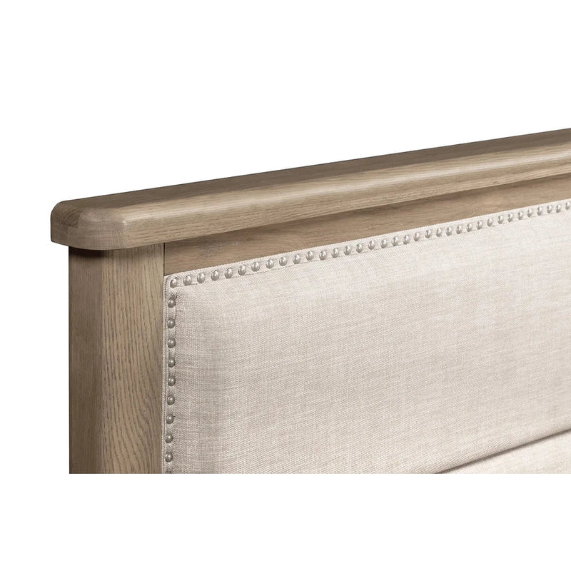 A close-up of the Aylesbury bedframe, showcasing its oak frame and light beige upholstered headboard, accented with a row of decorative silver nailhead trim along the edge.