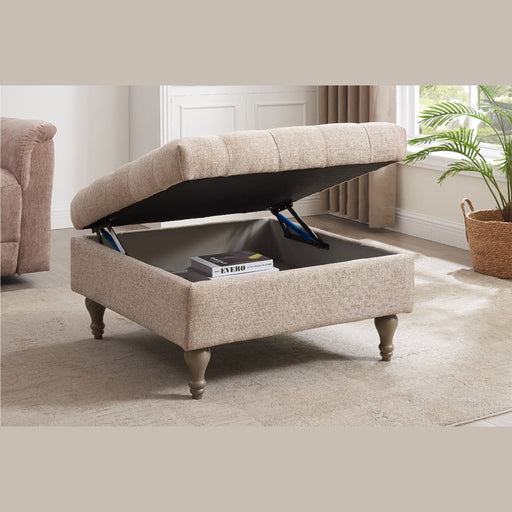 An Abbott Storage Footstool with a tufted lid is propped open to show books inside. This beige piece stands on wooden legs in a bright living room, paired with a beige rug and a nearby potted plant.
