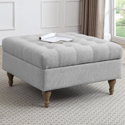 A square, light gray Abbott Storage Footstool with wooden legs is placed on a patterned rug in a bright living room. A magazine rests on top, with large windows and a plant visible in the background.