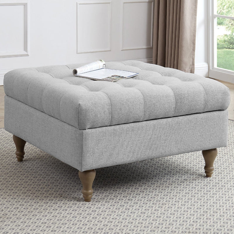 A square, light gray Abbott Storage Footstool with wooden legs is placed on a patterned rug in a bright living room. A magazine rests on top, with large windows and a plant visible in the background.