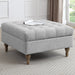A square, light gray Abbott Storage Footstool with wooden legs is placed on a patterned rug in a bright living room. A magazine rests on top, with large windows and a plant visible in the background.