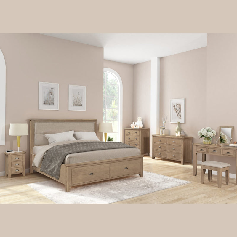 A modern bedroom featuring light wood furniture, including the Aylesbury bedframe, matching nightstands and dressers, a vanity, beige walls, wood floors, a large window, simple decor with framed art, and fresh flowers.