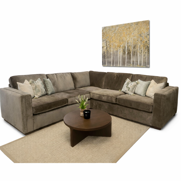 A cozy living room setup featuring the Ariya sofa for maximum comfort, assorted throw pillows, a round wooden coffee table on a beige rug, and wall art of tall trees with yellow leaves.
