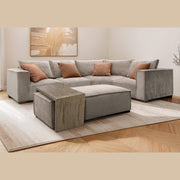 A modern living room featuring a gray modular sectional sofa, four brown patterned throw pillows, the Bodhi Ottoman + Coffee Table, a wooden side table, abstract wall art, and a light area rug on wood flooring.