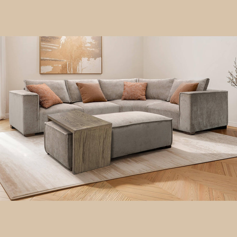 A modern living room featuring a gray modular sectional sofa, four brown patterned throw pillows, the Bodhi Ottoman + Coffee Table, a wooden side table, abstract wall art, and a light area rug on wood flooring.