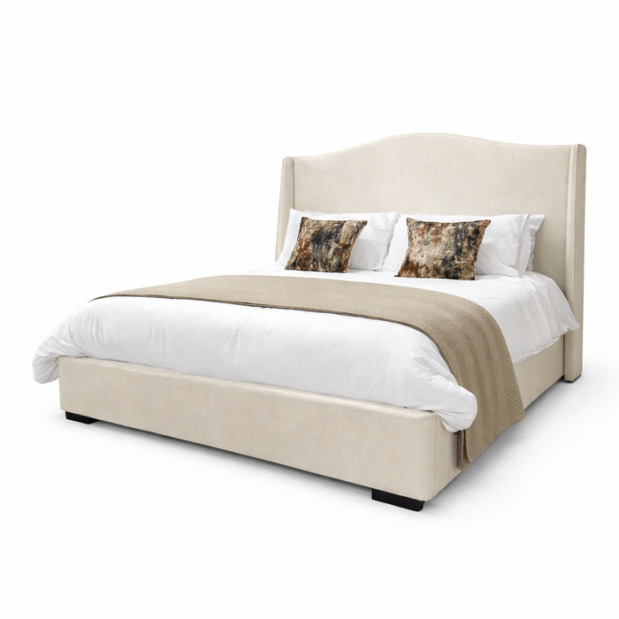 The Venice Bedframe is a modern luxury bed with a cream upholstered frame, tall curved headboard, white bedding, light brown throw blanket, and two abstract-patterned pillows, shown against a white background.
