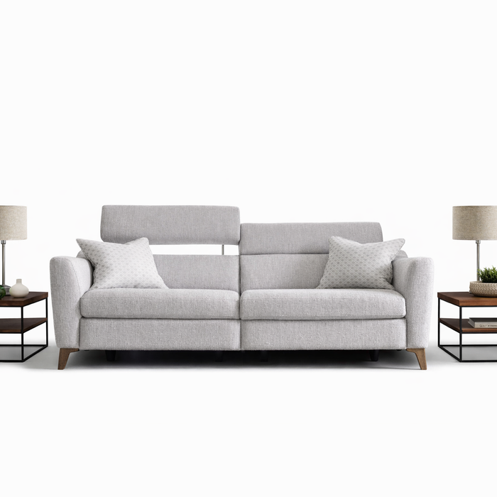 The Otis Sofa Range in light gray features adjustable headrests and two patterned cushions, accompanied by matching wooden side tables with lamps and decor, all displayed against a plain white background.