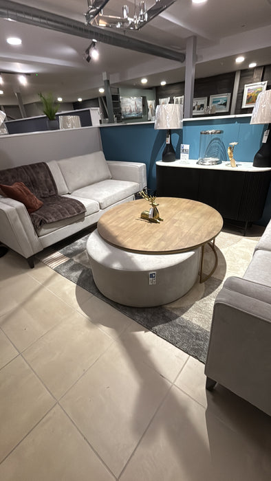 A modern living room showroom featuring two light gray sofas, the Todi Round Coffee Table & Stool Set, table lamps, and framed art on a blue accent wall, all arranged over a beige tiled floor.