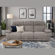 A modern living room features the gray Newbury Recliner Sofa with manual recline and easy-clean fabric, two throw pillows, a round wooden coffee table, a floor lamp, a potted plant, abstract art pieces, and large windows in the background.