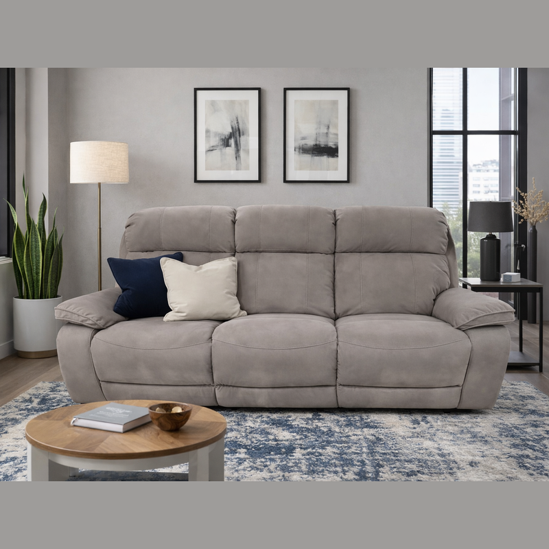 A modern living room features the gray Newbury Recliner Sofa with manual recline and easy-clean fabric, two throw pillows, a round wooden coffee table, a floor lamp, a potted plant, abstract art pieces, and large windows in the background.
